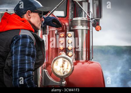 Ein Lkw-Fahrer in einem blauen karierten Hemd und einem roten Hoodie lehnt sich an einen klassischen roten Truck, der sich auf den Horizont konzentriert. Kühles Wetter trägt zur Atmosphäre bei Stockfoto
