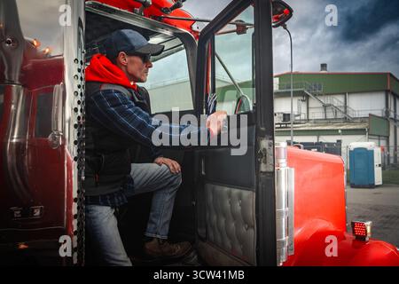 Ein LKW-Fahrer sitzt in einem roten LKW, bereit, seine Reise zu beginnen. Der Hintergrund zeigt ein Industriegebiet mit bewölktem Himmel. Stockfoto