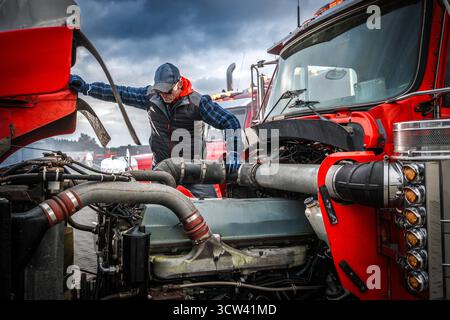 Ein Mechaniker mit einer Kappe arbeitet auf einem Lkw-Hof unter bewölktem Himmel an einem roten Lkw-Motor, um das Leistungsproblem des Fahrzeugs zu beheben. Stockfoto