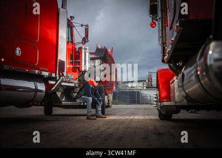 Zwei Lkw-Fahrer sitzen auf den Stufen eines großen roten Lkws und genießen eine Pause in einer industriellen Umgebung, während dunkle Wolken über ihnen hängen. Stockfoto