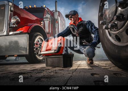 Ein Lkw-Fahrer kniet neben seinem großen Bohrfahrzeug und überprüft die Werkzeuge aus einer Kiste, während er am frühen Morgen unter bewölktem Himmel am Fahrzeug arbeitet. Stockfoto