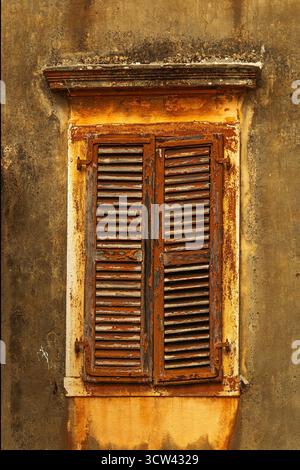 Ein Blick aus der Nähe auf alte Holzläden mit abblätternder Farbe und rostigen Scharnieren an einer strukturierten gelben Wand in Korfu Stadt. Stockfoto