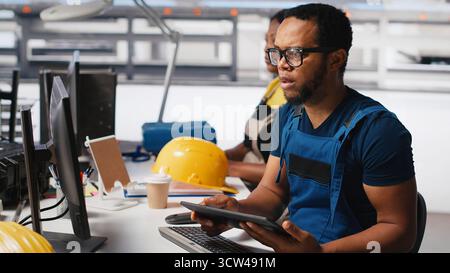Ingenieur untersucht fehlerhafte Systeme in Photovoltaik-Fabriken und vergleicht Parameter mit pc und Tablet. Schwarzer Mann inspiziert Solarpaneele, High-Tech. Kamera A.. Stockfoto