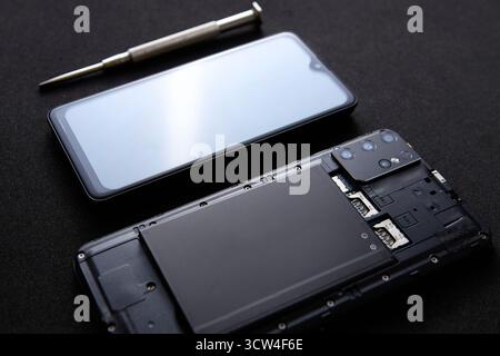 Ein Smartphone mit offenem Gehäuse und Schraubendreher, das auf einer reflektierenden schwarzen Oberfläche platziert ist. Konzeptbild für Technikwartungs- und Reparaturdienst Stockfoto