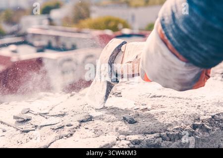 Mit Handschuhen trifft die Hand mit einem Hammer auf Beton, wodurch Staub und Fremdkörper während des Aufpralls fliegen. Die verschwommene urbane Kulisse deutet auf Arbeit im Freien hin Stockfoto