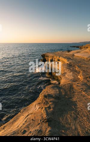 Wellen krachen sanft gegen die zerklüftete Küste, während die Sonne untergeht, und werfen warme Farben über die Landschaft und das Wasser am Abendhimmel. Stockfoto