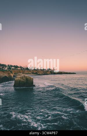 Der farbenfrohe Himmel bei Sonnenuntergang strahlt ein warmes Leuchten über der felsigen Küste aus. Sanfte Wellen schlagen gegen die Felsen in der Nähe des Ufers, wenn der Abend näher rückt. Stockfoto