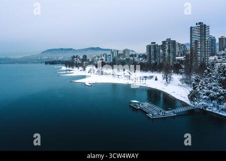 Eine ruhige Winterszene an einem See mit einer verschneiten Küste. Stadtgebäude erheben sich im Hintergrund und zeigen eine urbane Landschaft unter grauem Himmel. Stockfoto