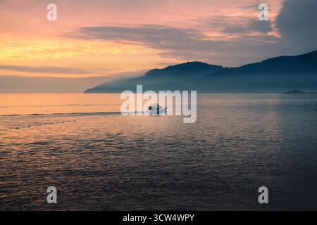 Ein kleines Boot fährt sanft über das ruhige Wasser, während ein lebhafter Sonnenuntergang den Himmel übermalt. Die nahe gelegenen Hügel bilden eine malerische Kulisse, die den Peac ergänzt Stockfoto