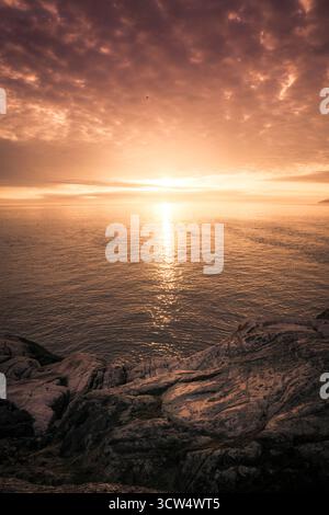 Ein atemberaubender Sonnenuntergang erfüllt den Himmel mit warmen Farben, während die Sonne über dem ruhigen Ozean untergeht. Wellen schlagen sanft gegen die zerklüfteten Felsen und erzeugen ein heiteres A Stockfoto