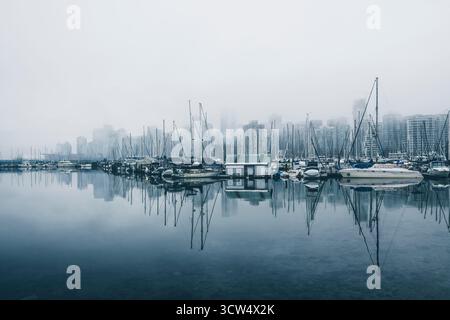 Die Boote liegen friedlich an einem Yachthafen und spiegeln sich im stillen Wasser. Hohe Gebäude ragen im Nebel und schaffen eine ruhige Atmosphäre an diesem kühlen Morgen Stockfoto
