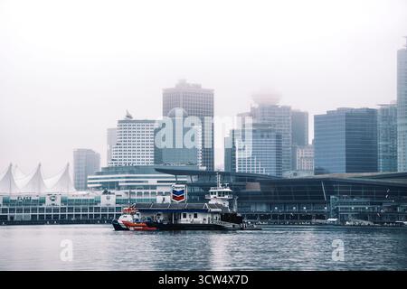 Nebel bedeckt die Skyline der Stadt, während Boote durch das ruhige Wasser fahren. Wolkenkratzer erheben sich im Hintergrund und schaffen schon früh eine ruhige urbane Szene. Stockfoto