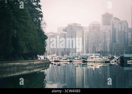Hohe Gebäude erheben sich im Nebel über einem Hafen voller Boote. Das stille Wasser spiegelt das Stadtbild wider und schafft in den frühen Jahren eine ruhige Stadtlandschaft Stockfoto