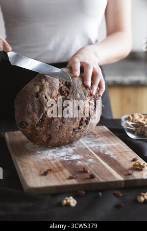 Frau, die ein rustikales Roggensauerteigbrot mit Walnüssen und Rosinen auf einem Holzbrett aufschneidet und seinen handwerklichen, hausgemachten Stil zeigt. Stockfoto