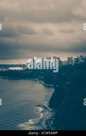 Die Skyline einer geschäftigen Stadt steht hoch vor dunklen Wolken, darunter ist eine ruhige Uferpromenade zu sehen. Wellen schlagen sanft am Rand und schaffen Frieden Stockfoto
