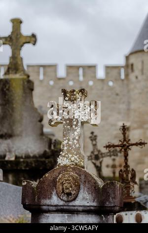 Ein altes Steinkreuz bedeckt mit Flechten und ein Basrelief von Jesus Christus auf einem alten Friedhof, neben einer mittelalterlichen Festungsmauer im Hintergrund. Stockfoto