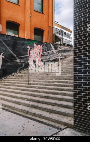 Blick auf eine Betontreppe im Freien, flankiert von einem orangen Gebäude und einer dunklen Ziegelmauer, mit einem ausdrucksstarken Wandgemälde mit Katzencharakter. Ideal für urba Stockfoto