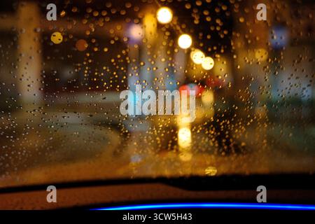 Regen fällt nachts auf die Windschutzscheibe des Autos mit Stadtlichtern Bokeh, Blick aus dem Inneren des Fahrzeugs während der regnerischen Abendfahrt. Stockfoto