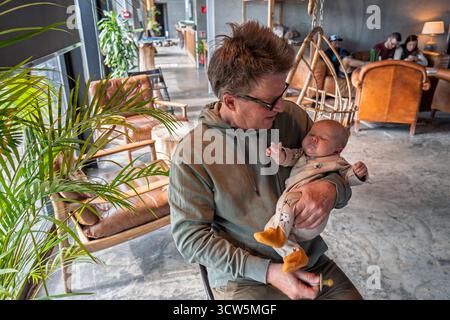 Großvater, der ein Neugeborenes in einem gemütlichen Café hüllt – ein zärtlicher, liebevoller Moment über Generationen hinweg. Stockfoto