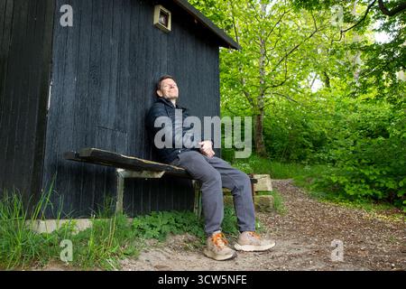 Mann im friedlichen Moment auf Bank im Wald genießt die Natur, Mann lehnt sich an Holzschuppen und glücklich im Naturmoment Stockfoto