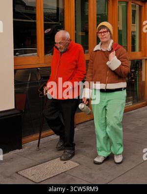 Ein älterer Mann, der sich auf einen Stock stützt und in einem leuchtend orangefarbenen Fleece gekleidet ist, geht an der Seite einer jüngeren Frau in einem leuchtend mintgrünen Outfit und einer gelben Beanie. Stockfoto