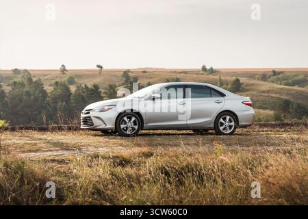 Silver Toyota Camry in hügeliger Gegend. Seitenansicht der japanischen Limousine auf trockener Wiese, Hügel mit Bäumen im Hintergrund. Stockfoto