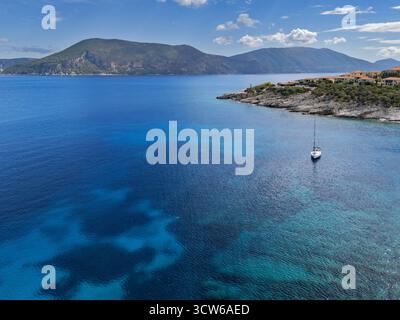 Aus der Vogelperspektive auf einem einsamen Segelboot, das auf dem kristallklaren türkisfarbenen Wasser gleitet, eingerahmt von einer felsigen Küste und entfernten sanften Hügeln, Fiskardo, Kefalonia, Griechenland. Stockfoto