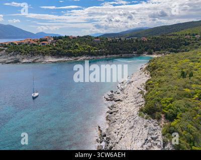 Aus der Vogelperspektive auf türkisfarbenes Wasser treffen auf felsige Küsten und üppige grüne Hügel, mit einem einsamen Segelboot, das friedlich gleitet, Fiskardo, Kefalonia, Griechenland. Stockfoto