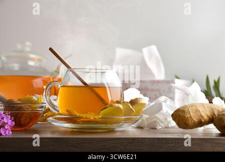 Hausmittel gegen Erkältungen und Stauungen mit einer natürlichen Infusion aus Ingwerwurzel und Honig in einer Glasschale auf einem Holztisch mit gebrauchten Taschentüchern und einem Stockfoto