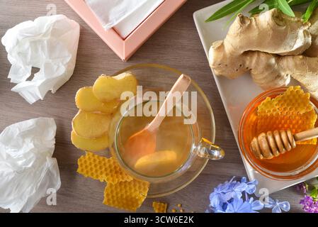 Hausmittel gegen Erkältungen und Stauungen mit einer natürlichen Infusion aus Ingwerwurzel und Honig in einer Glasschale auf einem Holztisch mit gebrauchten Taschentüchern und einem Stockfoto