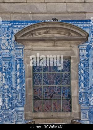 Barockes Fenster und Azulejos in Porto, Portugal - Nahaufnahme eines Kirchenfensters mit Buntglasfenster, das von barockem Stein eingerahmt und von traditionellem Blau umgeben ist Stockfoto