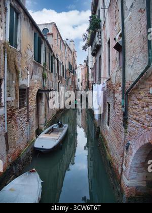 Venedig versteckter Kanal mit Boot und Himmelsspiegelung - Moody, stimmungsvolle Aufnahme eines engen venezianischen Kanals mit leerem Boot, altes Backsteingebäude Stockfoto