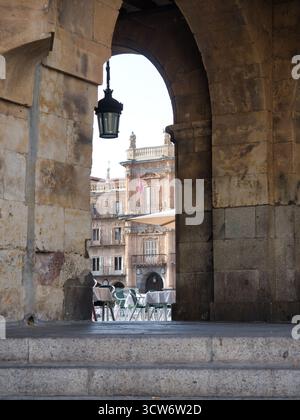 Bogengang am Salamanca Balkon - Blick durch einen dunklen Bogen auf historische goldene Steinarchitektur und Café-Terrasse. Stockfoto