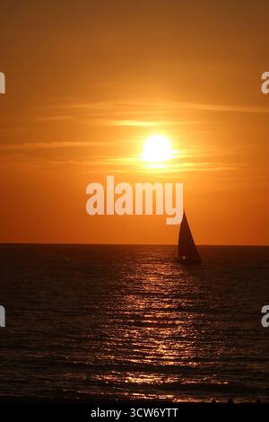 Sailboat Silhouette am Golden Sunset - Ein einsames Segelboot, das vor einem leuchtend orangen Himmel steht, während die Sonne über dem ruhigen Ozean untergeht und einen schönen Blick schafft Stockfoto