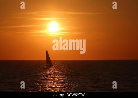 Sailboat Silhouette am Golden Sunset - Ein einsames Segelboot, das vor einem leuchtend orangen Himmel steht, während die Sonne über dem ruhigen Ozean untergeht und einen schönen Blick schafft Stockfoto