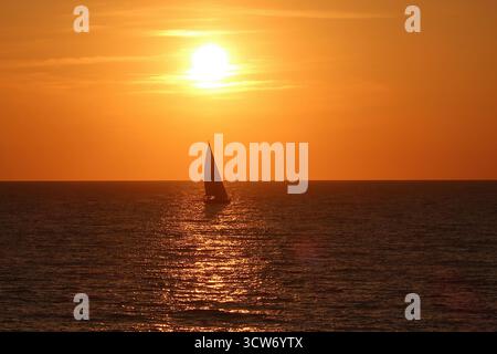 Sailboat Silhouette am Golden Sunset - Ein einsames Segelboot, das vor einem leuchtend orangen Himmel steht, während die Sonne über dem ruhigen Ozean untergeht und einen schönen Blick schafft Stockfoto