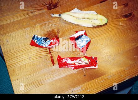 Bananenschale und Schokoladenbananenverpackung liegen auf einem Holztisch, scharf beleuchtet durch Blitz. Alltagsgegenstände mit rauen, lebendigen Kontrasten. Stockfoto
