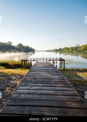 Hölzerner Pier auf dem See (symmetrische abnehmende perspektivische Sicht). Herbstvormittagsszene mit Nebel über dem Wasser, Bäumen und Himmel, die sich in der Seeoberfläche spiegeln Stockfoto