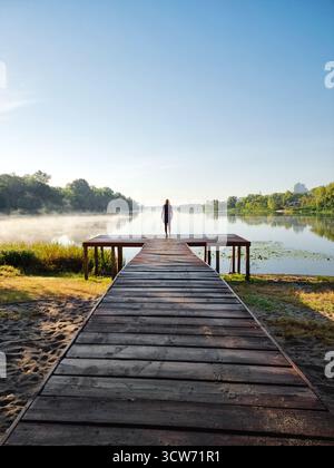 Silhouette einer Person auf hölzernem Pier. Herbstvormittagsszene mit Nebel über dem See, Bäumen und Himmel, die sich in spiegelförmiger Seenoberfläche spiegeln. Stockfoto