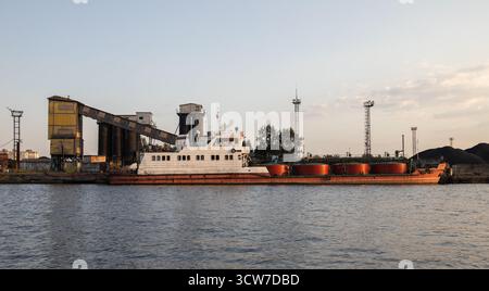 Ein altes Tankschiff sitzt an einem geschäftigen Industriehafen mit großen Förderanlagen, Silos und Kränen entlang des Kais. Die Szene vermittelt Transport, Verladung und m Stockfoto