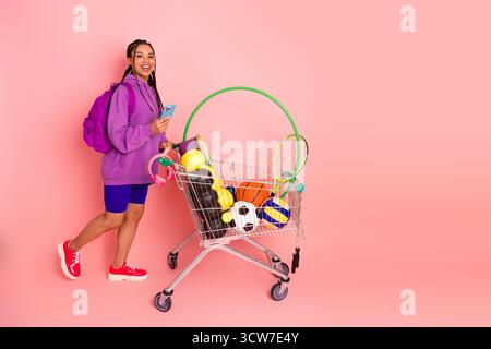 Sportliche Frau mit Rucksack, die einen Einkaufswagen mit Fitnessgeräten schiebt, vor rosafarbenem Hintergrund Stockfoto