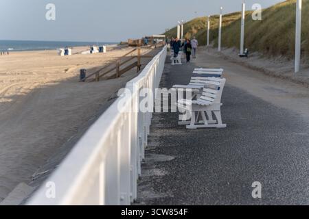 Promenade mit Bänken. Weiße Bänke säumen die Strandpromenade mit Blick auf den Sandstrand. Die Leute laufen im Hintergrund unter klarem Himmel. Stockfoto