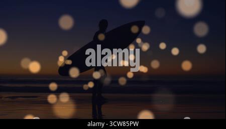 Wandelnder Surfer mit Surfbrett auf nassem Sand in der Abenddämmerung, mit Bokeh-Kugeln, die den Himmel reflektieren. Strand, Silhouette, Sonnenuntergang, Reflexion, Ruhe, Stockfoto