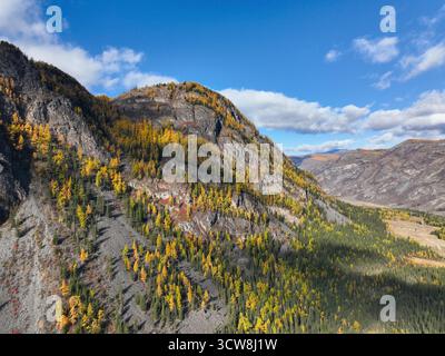 Aus nächster Nähe sehen Sie einen dramatischen, zerklüfteten Berghang mit gelben und grünen Bäumen unter einem hellblauen, bewölkten Himmel Stockfoto