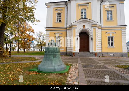 Auf dem Rasen eines Klosters ist eine massive, verwitterte Bronzeglocke ausgestellt, mit dem kunstvollen Eingang zu einem historischen gelben Gebäude im Hintergrund Stockfoto