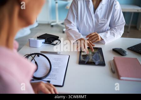 Kaukasische Frau mittleren Alters, die gegenüber jungen Erwachsenen kaukasische Ärztin sitzt, während der Krebsberatung in der Arztpraxis eine Röntgenaufnahme der Brust auf einem digitalen Tablet überprüft Stockfoto
