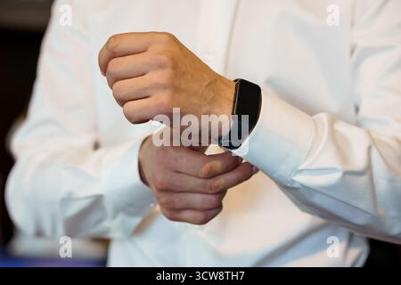 Eine Nahaufnahme zeigt einen Mann, der den schwarzen Riemen einer modernen Smartwatch am Handgelenk befestigt, während er ein weißes Hemd trägt, was auf ein Geschäft hindeutet Stockfoto