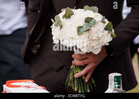 Eine Nahaufnahme zeigt einen Bräutigam, der eine weiße Nelkenboutonniere mit Eukalyptusblättern hält, ergänzt durch seinen braunen Anzug während einer Hochzeitsfeier Stockfoto