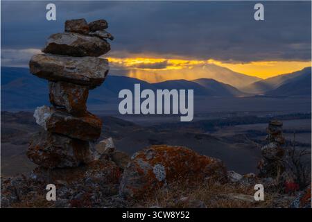 Gestapelte Felsen im Vordergrund überblicken das weite Tal, während die Sonne durch Wolken bricht und goldene Strahlen auf fernen Bergen wirft Stockfoto
