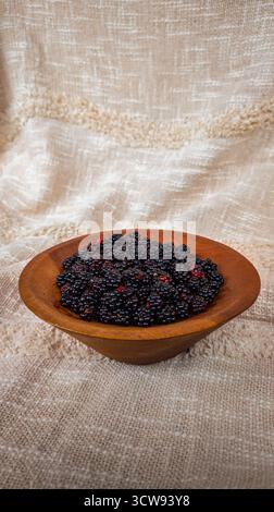 Ein rustikales Stillleben mit einer Holzschale gefüllt mit frischen Brombeeren auf beigefarbenem texturiertem Stoff. Stockfoto